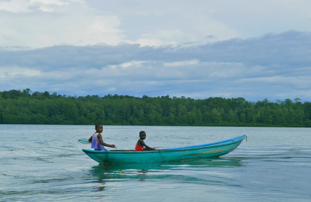 Dos niños navegan en una canoa de madera turquesa sobre aguas tranquilas frente al manglar