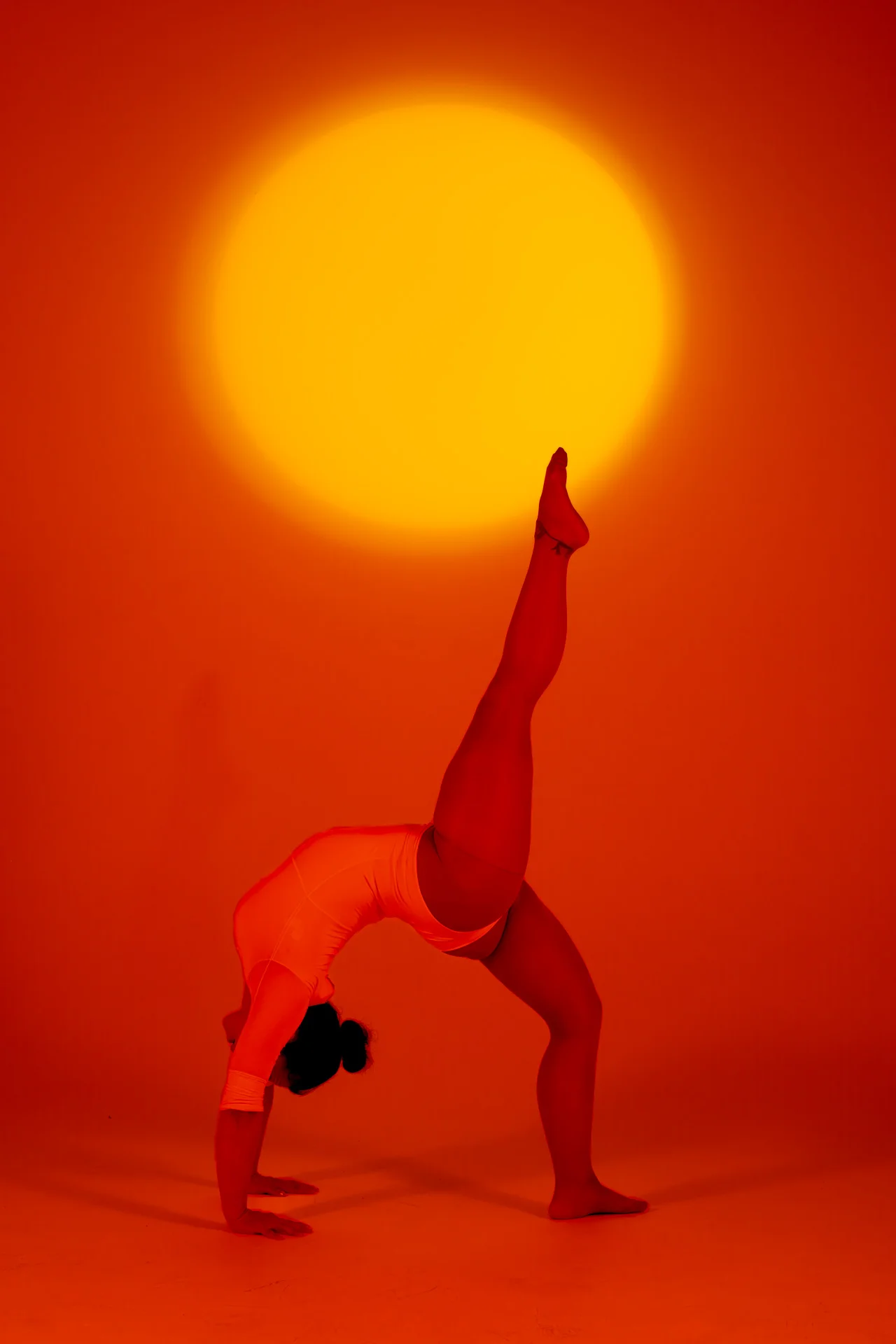 Mujer en una flexión hacia atrás con una pierna extendida hacia arriba bajo luz naranja cálida con un sol de fondo