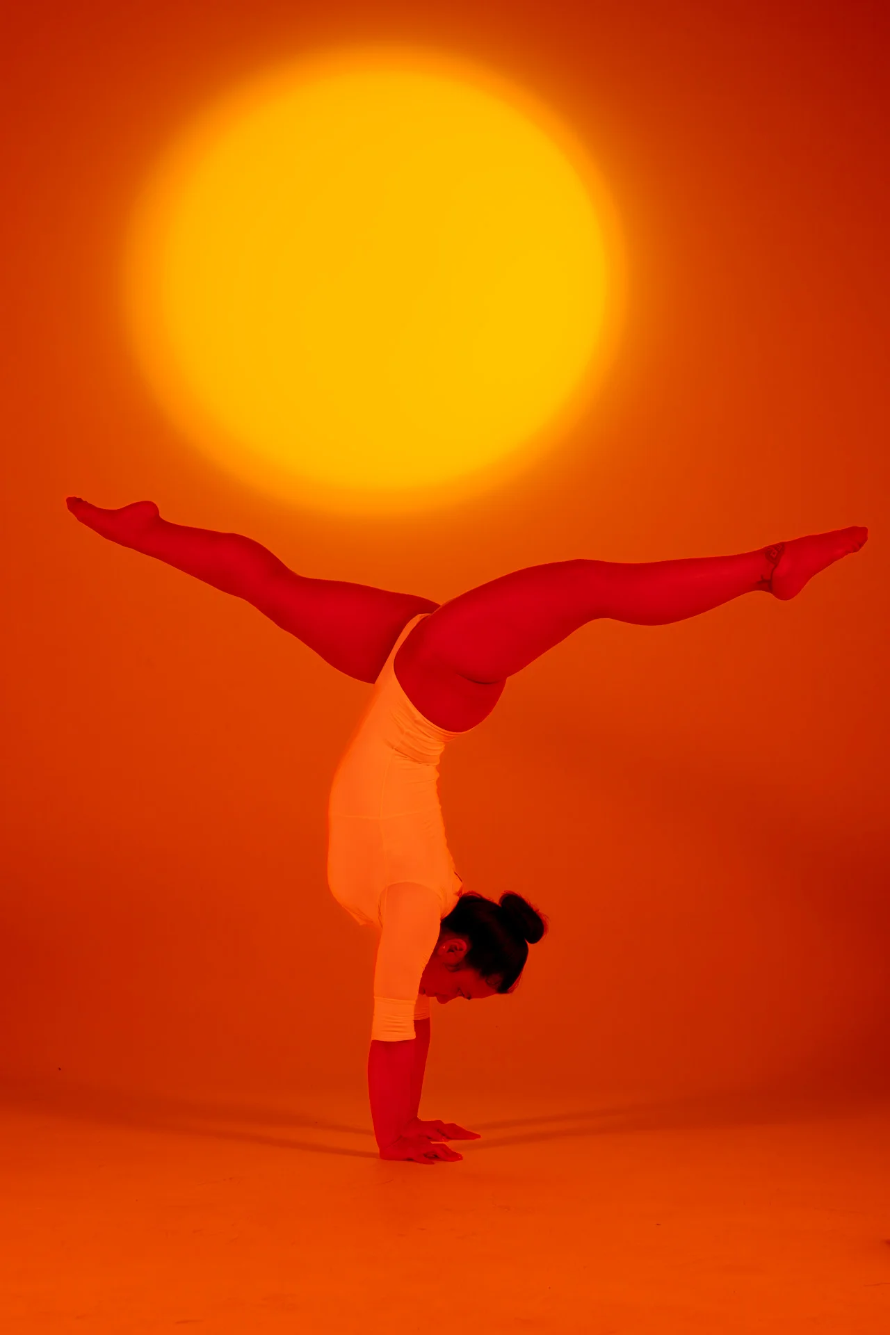 Mujer realizando una parada de manos con las piernas abiertas en horizontal bajo luz naranja cálida con un sol de fondo