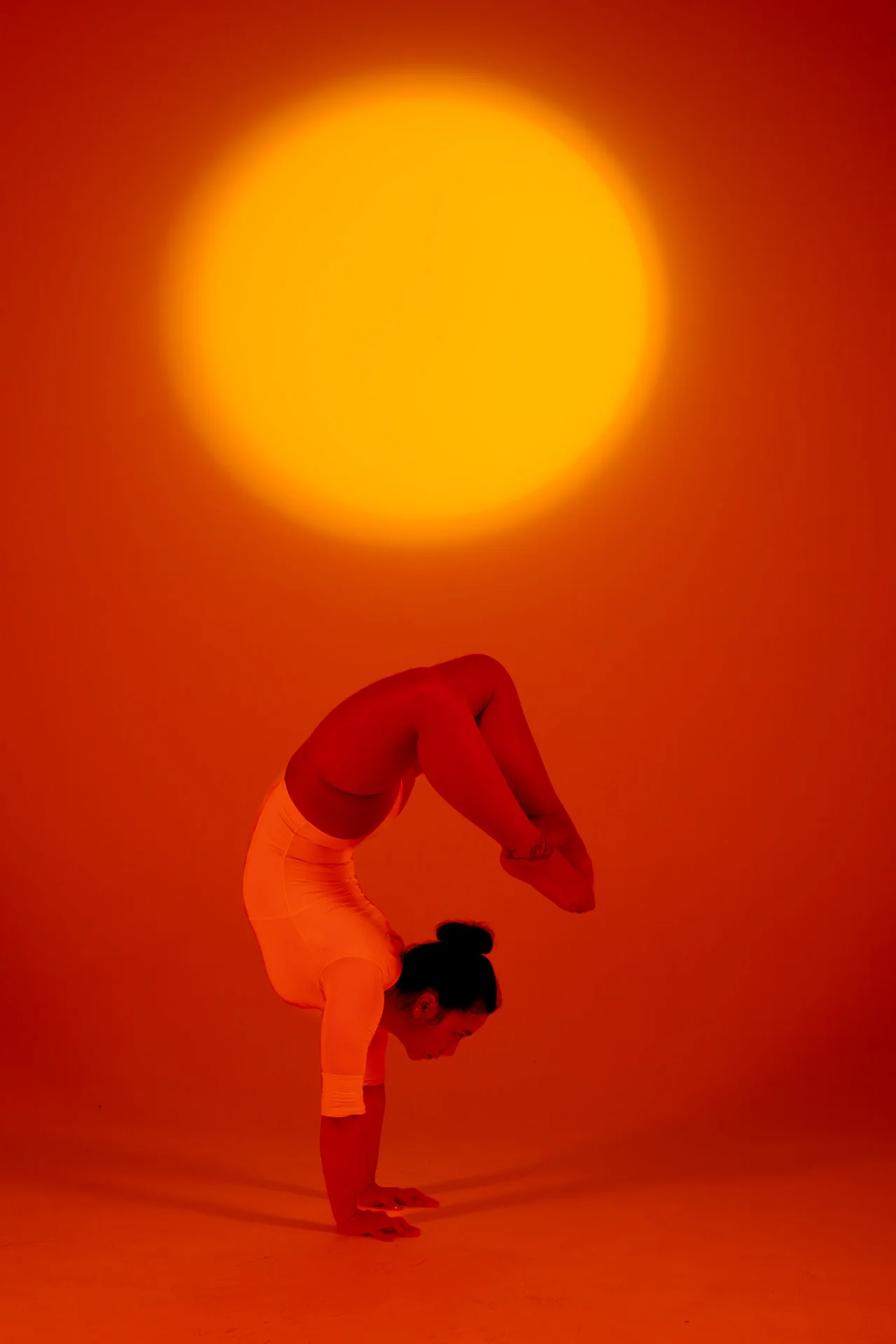 Mujer en una pose de flexión tipo escorpión bajo luz naranja cálida con un sol de fondo